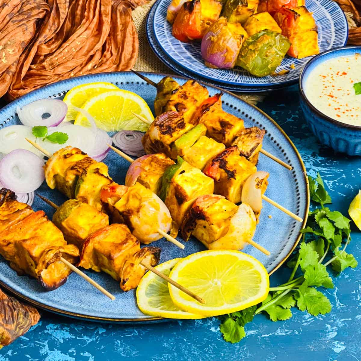 Paneer tikka threaded in skewers placed in a blue plate and served with mint chutney.