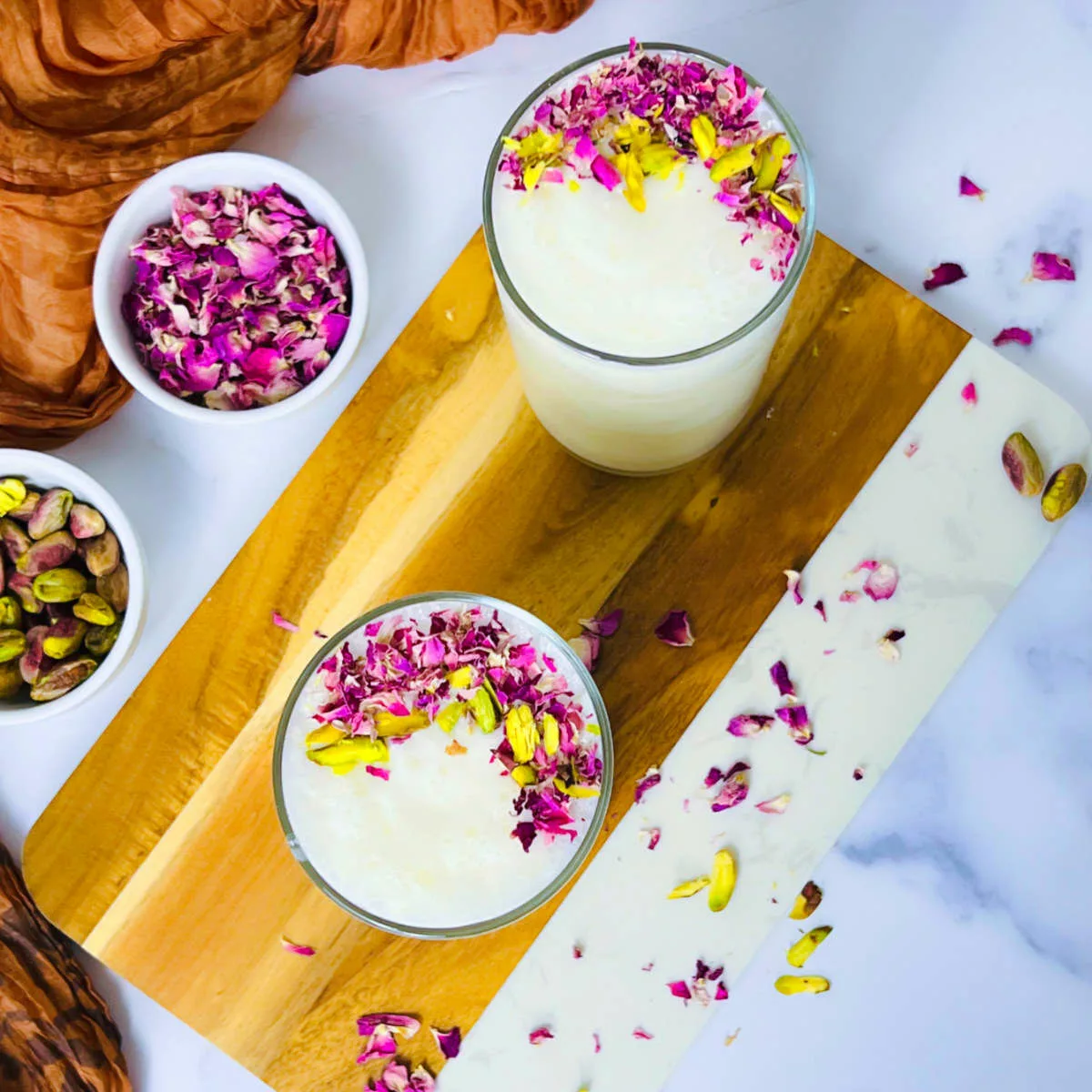 Sweet lassi placed on a wooden board with rose petals and pistachio in the background.