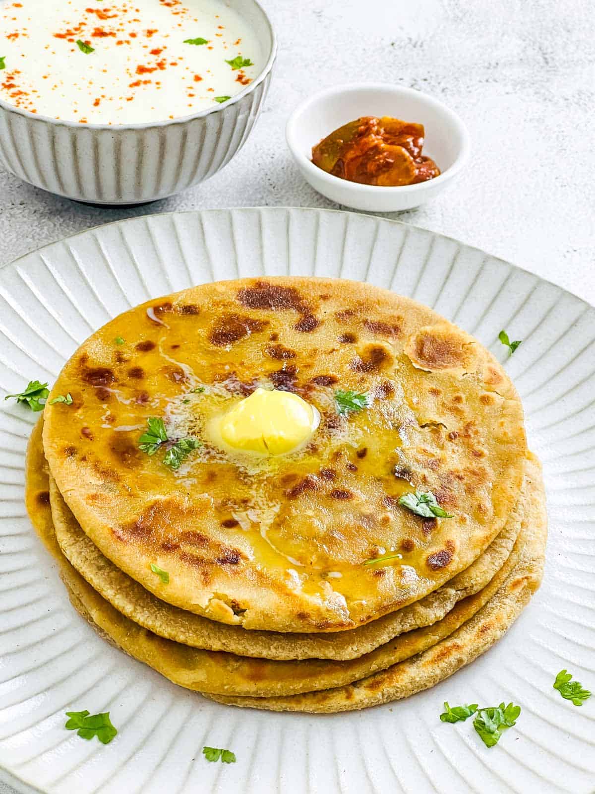 A stack of aloo paratha with butter melting on top placed on a white plate.