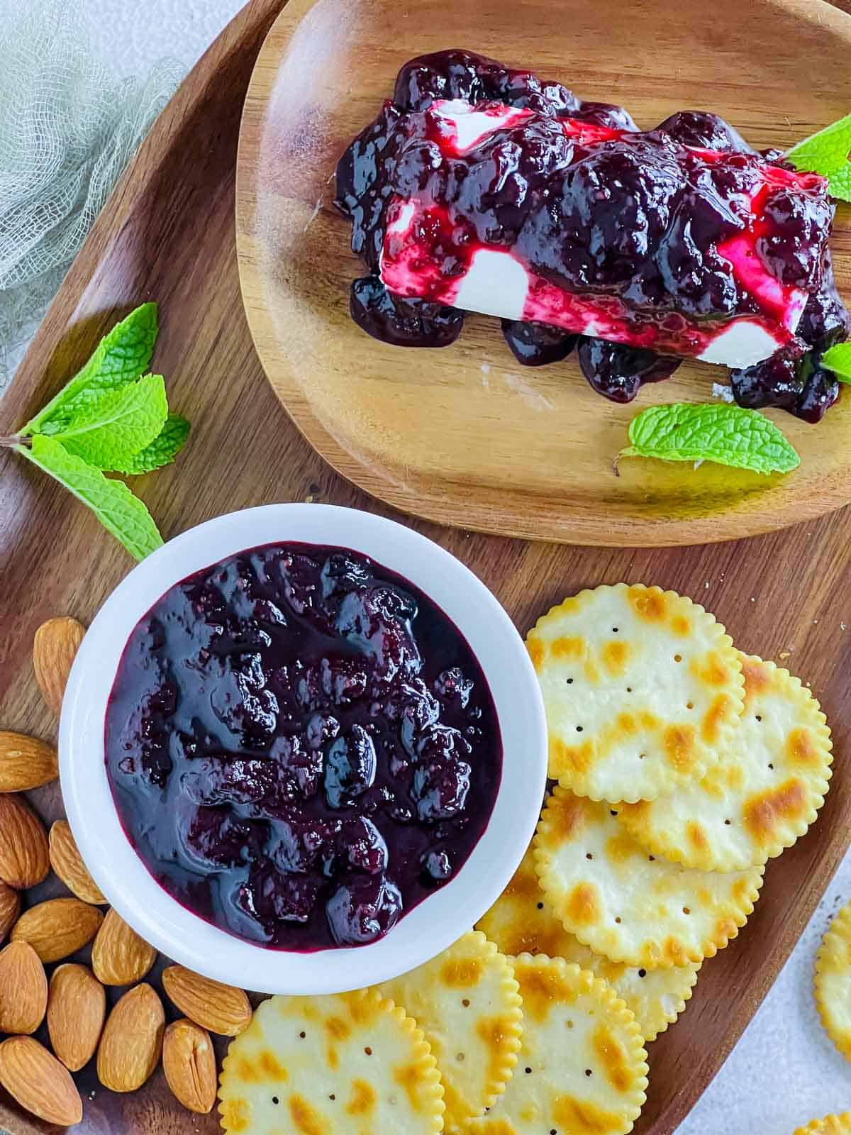 Blueberry chutney in a white bowl placed on a wooden board with crackers, nuts, and goat cheese.