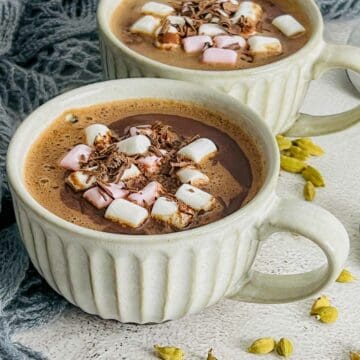 Two mugs of cardamom hot chocolate placed on a white surface.