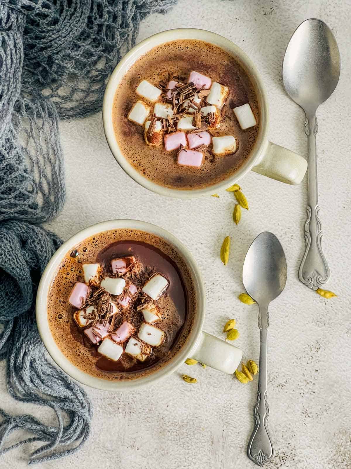Two mugs of cardamom  hot chocolate topped with marshmallow and placed on a white surface.