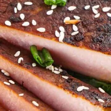 Close-up of sliced grilled pork belly garnished with sesame seeds and green chilies on a bed of kale.