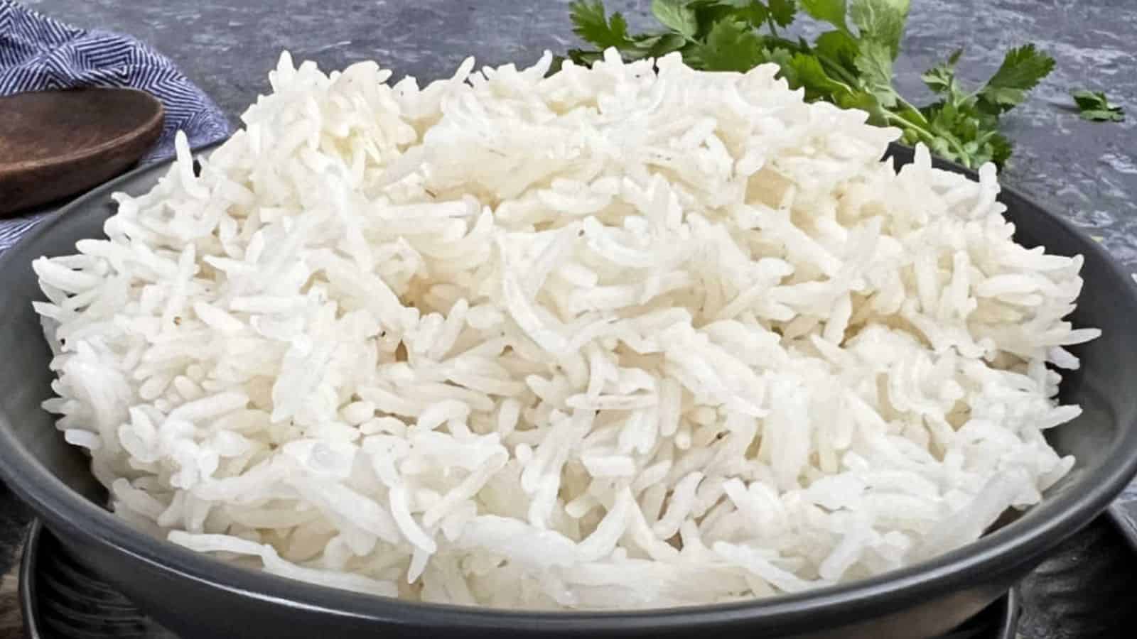 A bowl of cooked Basmati Rice on a gray surface, garnished with parsley, with a wooden spoon nearby.