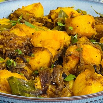 A bowl of Bombay Potatoes garnished with herbs, featuring golden potatoes and onions in a rich, spiced sauce.