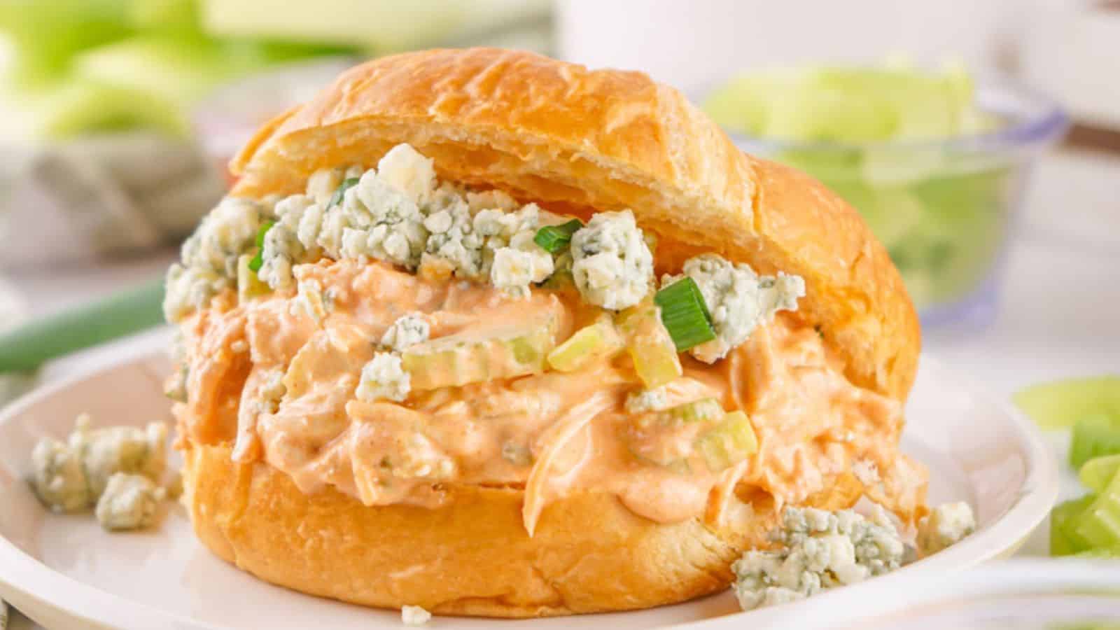 A croissant sandwich filled with buffalo chicken and blue cheese, served on a white plate with celery sticks in the background.