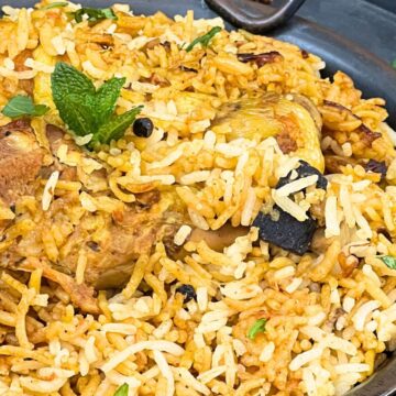 A close-up image of chicken biryani in a copper dish, garnished with mint and coriander leaves.