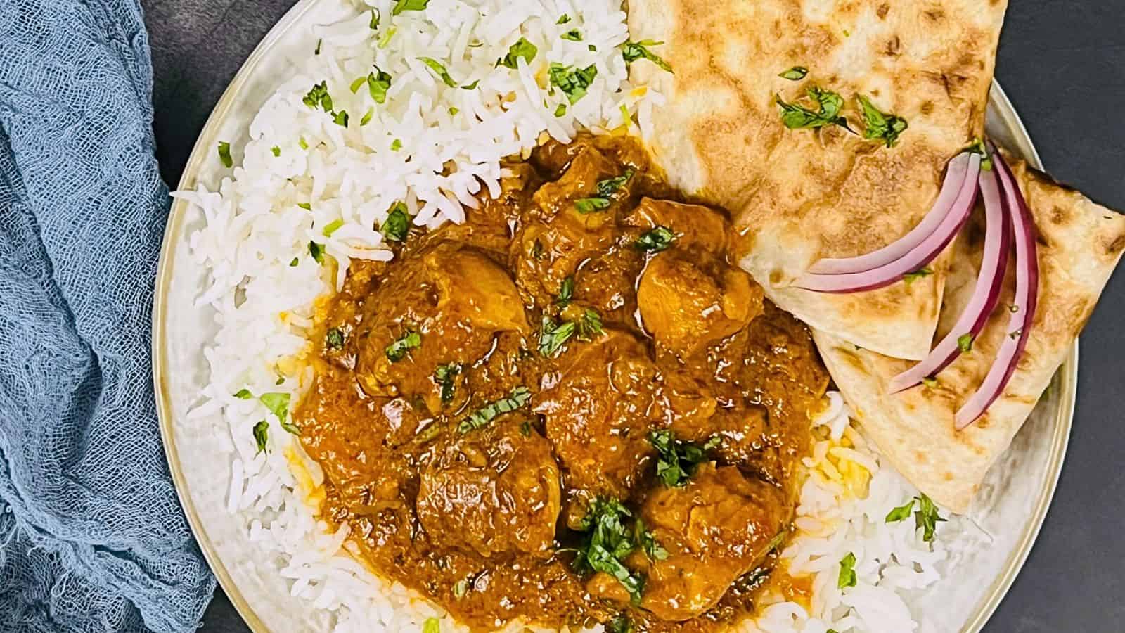A plate of Chicken Masala with two pieces of naan and basmati rice garnished with herbs, served with sliced onions on the side.