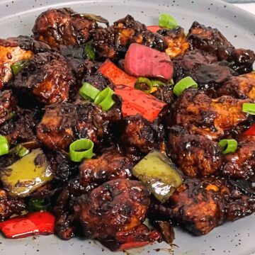 A plate of Chilli Chicken with chunks of red peppers, drizzled in a dark sauce, garnished with green onions.