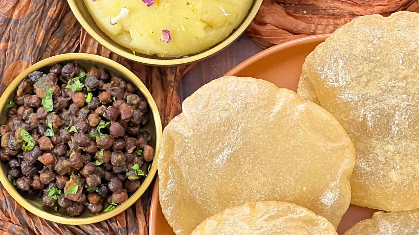 Image of Halwa Puri (Brunch/Chana) an indian meal featuring puris (fried bread), a bowl of black chickpeas, and mashed potatoes garnished with herbs.