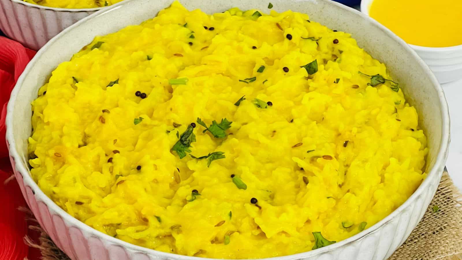A bowl of yellow rice garnished with herbs and spices, placed on a red mat with a glass of orange juice in the background.