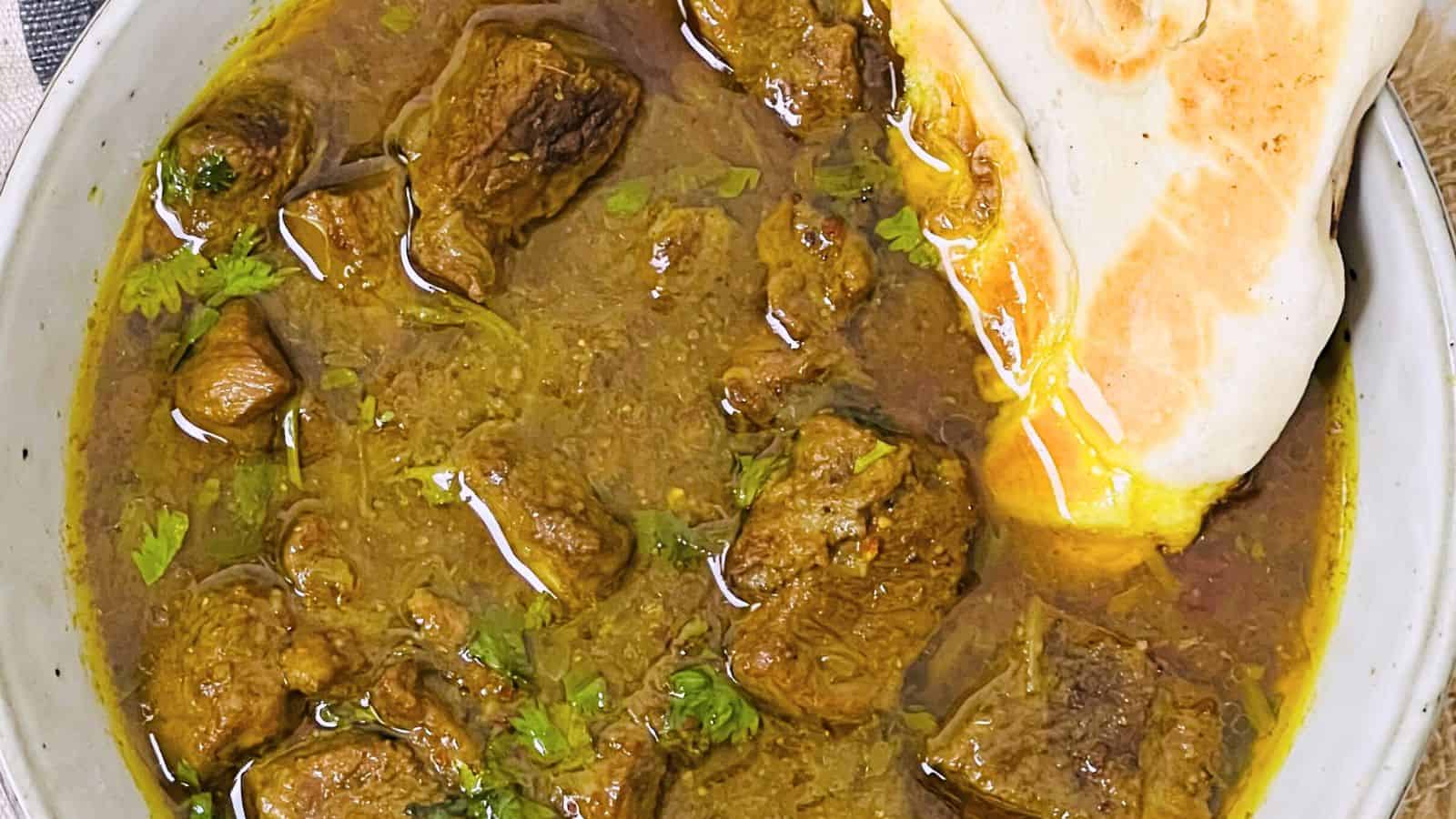 A bowl of rich beef curry garnished with cilantro, accompanied by a piece of naan bread partially dipped in the sauce.