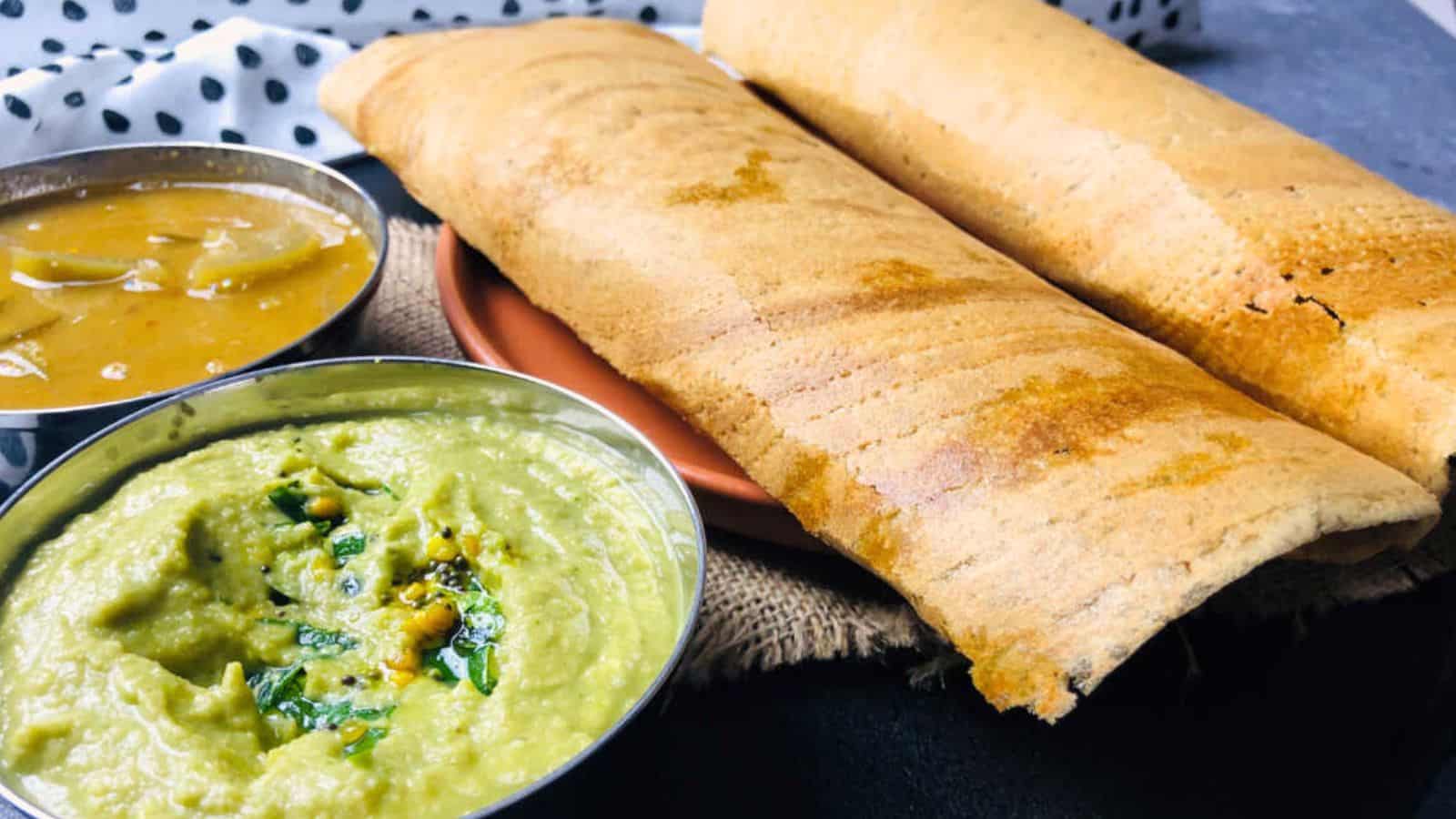 Two dosas served with coconut chutney and sambar in small bowls, placed on a rustic fabric surface.