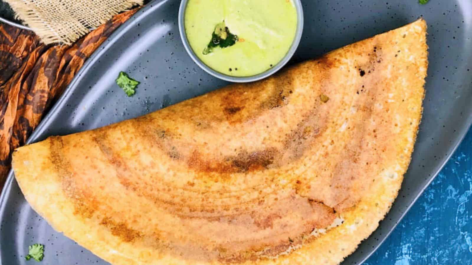 A folded, Mysore Masala Dosa on a dark plate served with a side of green chutney.