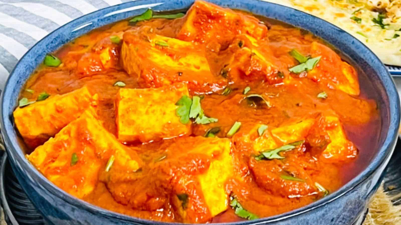 A bowl of Paneer Butter Masala with vibrant orange sauce garnished with herbs, served with naan bread in the background.