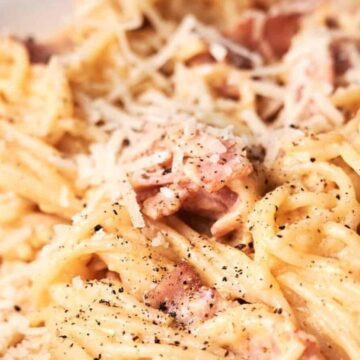 A close-up of creamy spaghetti carbonara sprinkled with grated cheese and black pepper, served on a white plate.