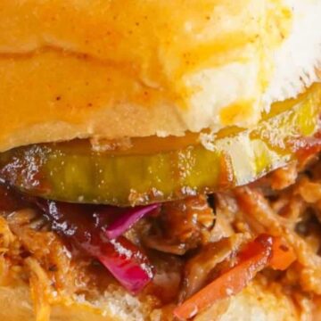 Close-up view of a pulled pork sandwich with pickles and red cabbage on a bun, displaying a savory and colorful meal.