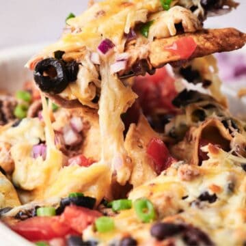 A bowl of taco casserole topped with melted cheese, black olives, tomatoes, and green onions, with a creamy dip in the background.