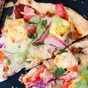 Slices of Tandori Paneer Naan Pizza with tofu, red onions, tomatoes, and drizzled with green sauce on a black surface.