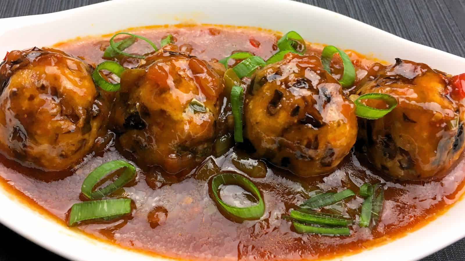 Four Manchurian meatballs in tomato sauce garnished with sliced green onions, served in a white oblong dish.