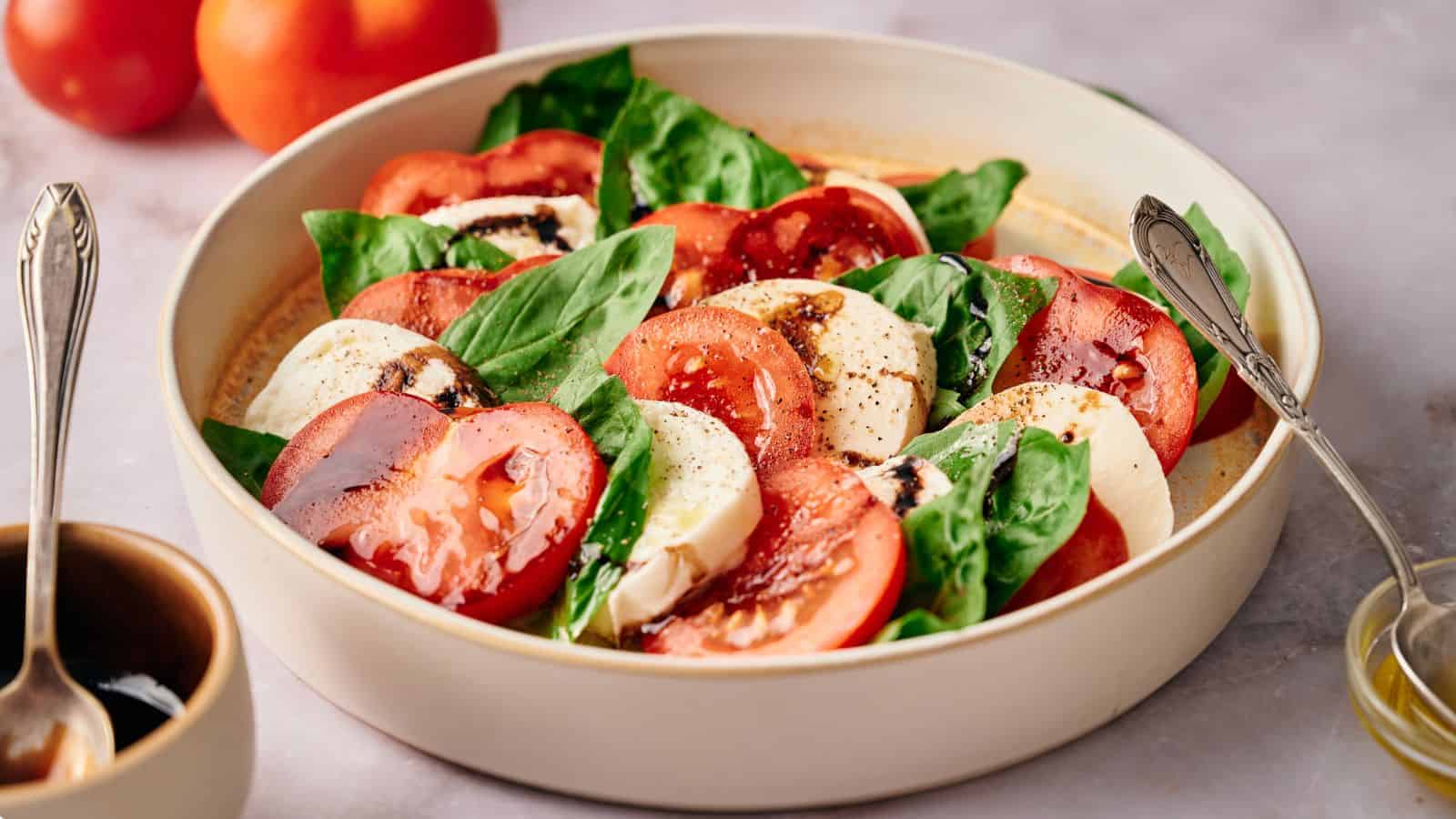 A bowl contains a Caprese salad made of fresh tomato slices, mozzarella, and basil leaves, drizzled with balsamic reduction and olive oil. Two tomatoes and a small bowl are in the background.