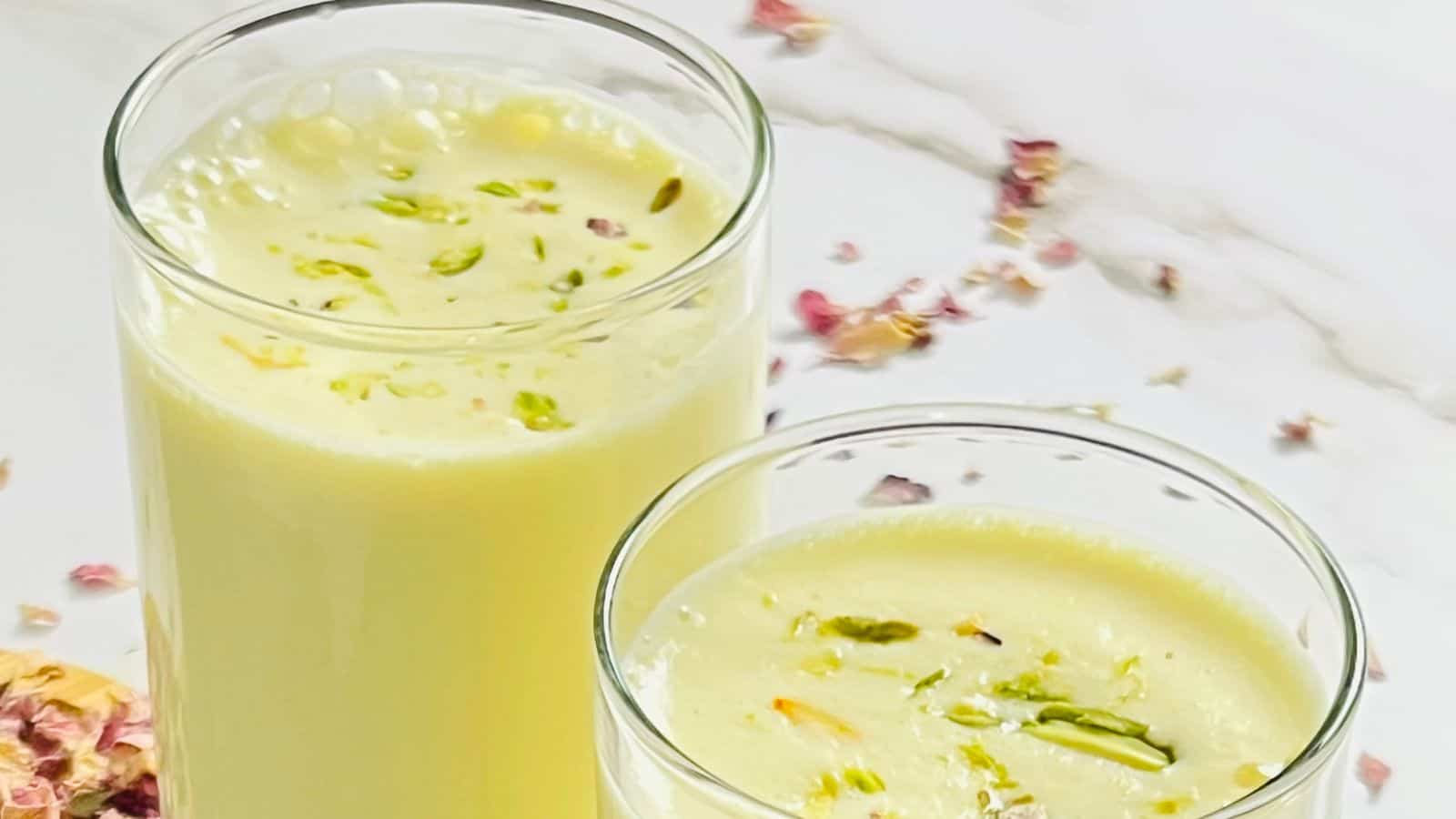 Two glasses of a creamy yellow drink topped with chopped pistachios and surrounded by flower petals on a white surface.