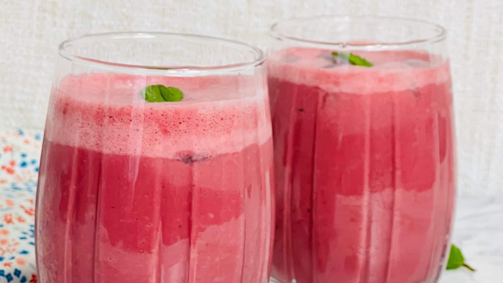 Two tall glasses filled with a frothy pink beverage, garnished with small mint leaves, placed on a light-colored surface with a patterned cloth nearby.