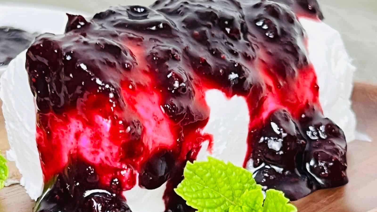 A close-up view of a dessert featuring white cheesecake topped with a rich, glossy berry sauce, garnished with fresh green mint leaves.