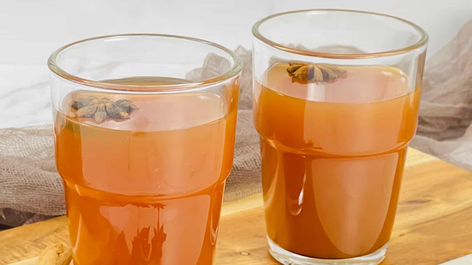 Two glasses of amber-colored liquid containing star anise flowers sit on a wooden surface, with a light fabric in the background.