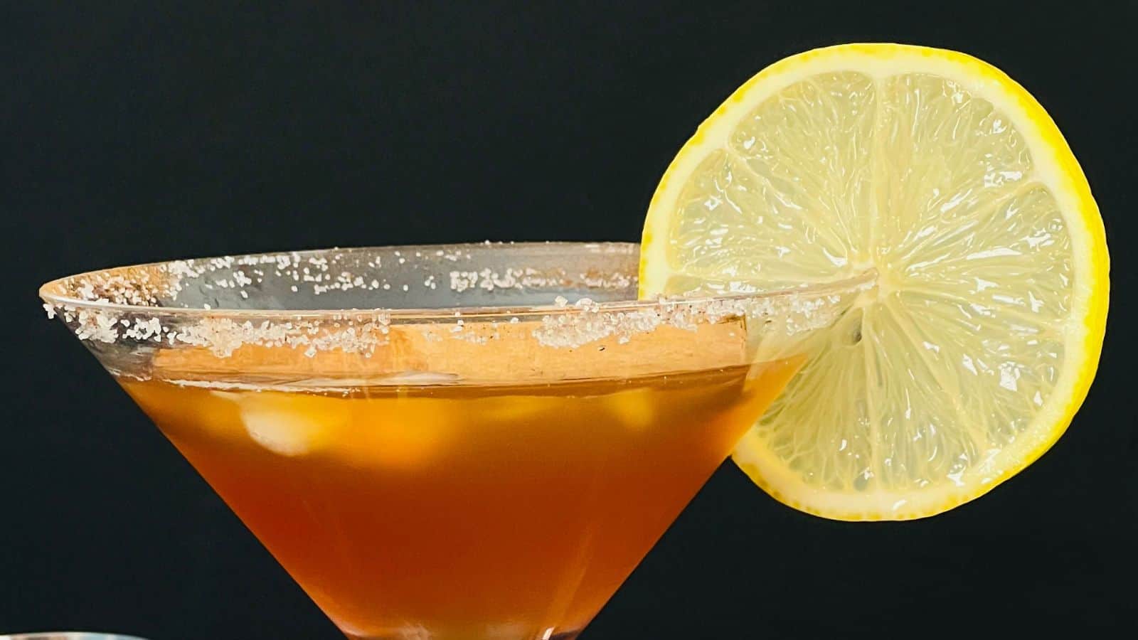 A martini glass with a salted rim, filled with an amber-colored drink, is garnished with a lemon slice.