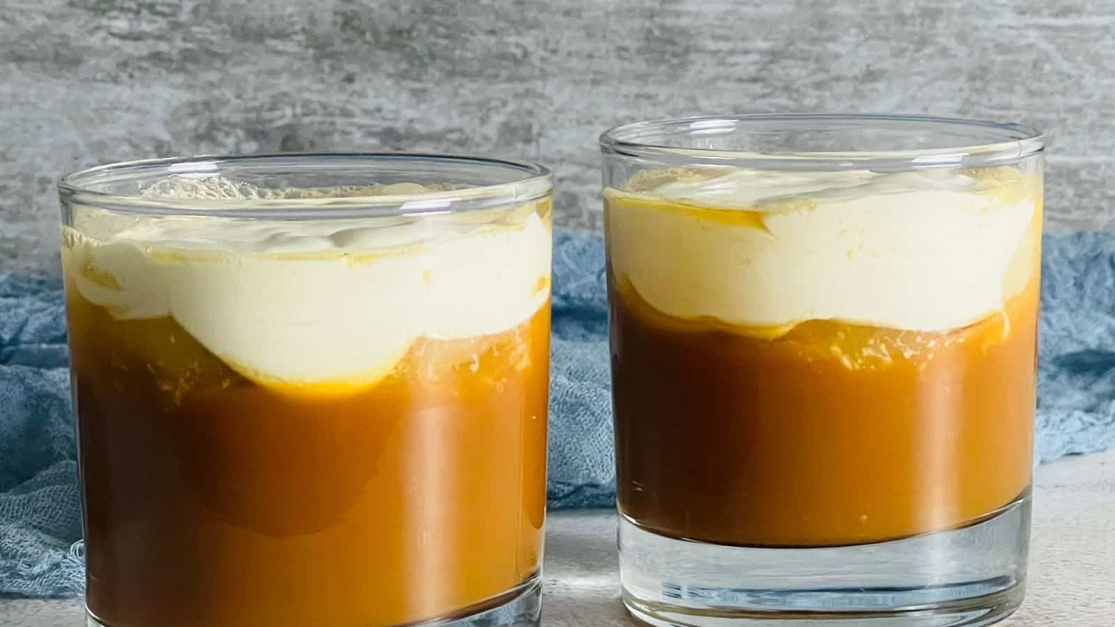 Two glass cups are filled with a layered dessert, featuring a golden caramel base topped with a creamy white layer, placed on a textured gray surface with a blue cloth in the background.