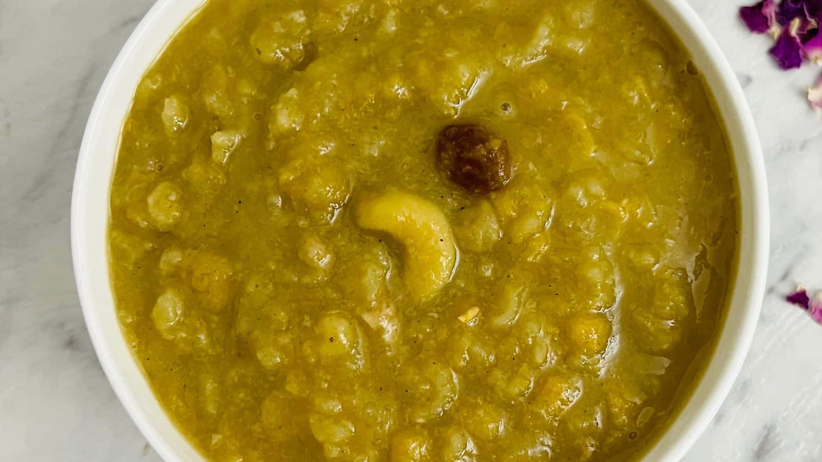 A bowl of thick, yellow-colored porridge with visible chunks, including one cashew and a raisin, on a white surface.