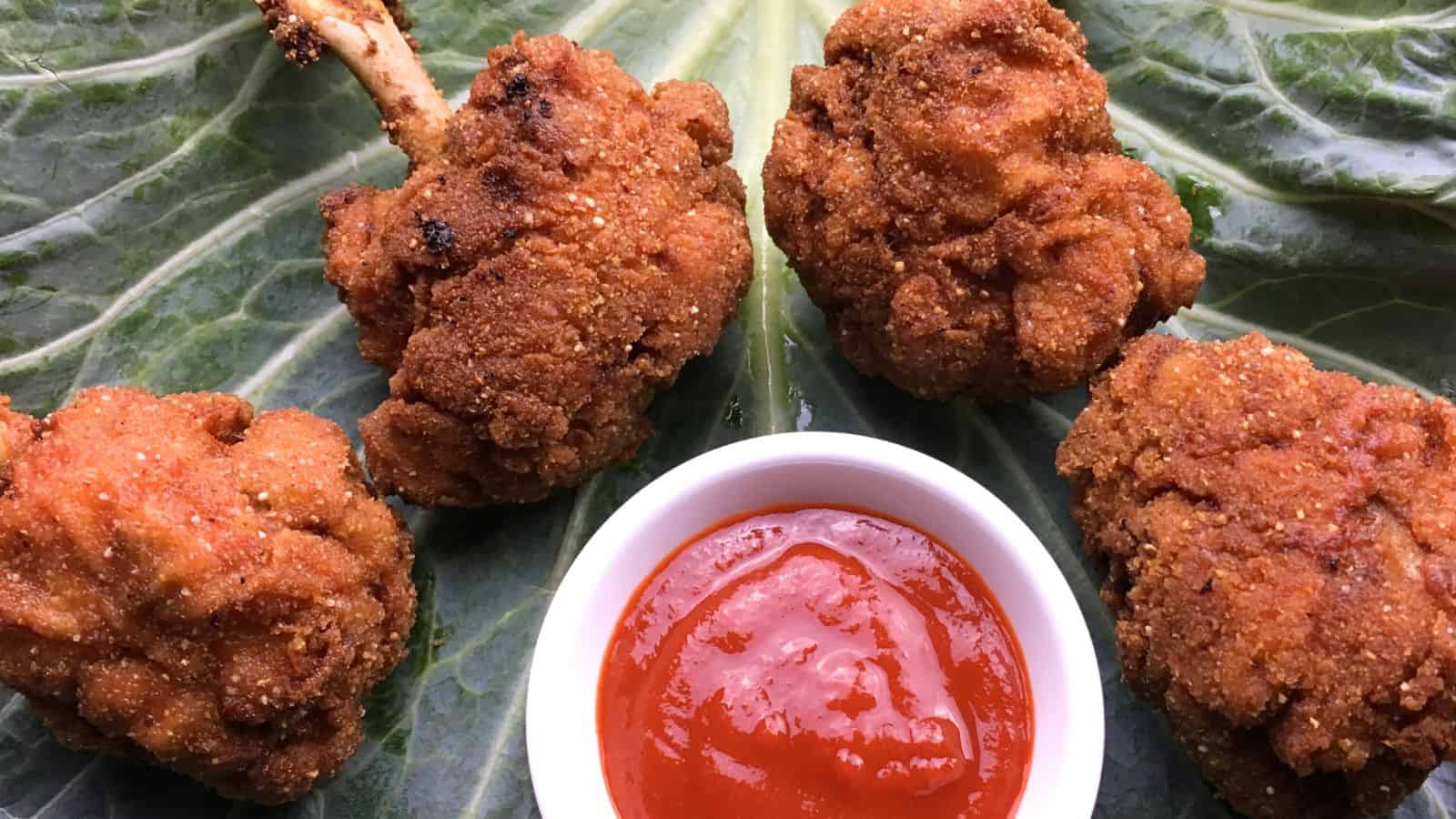 Four pieces of breaded, fried chicken arranged on green leaves with a small white bowl of red dipping sauce in front of them.