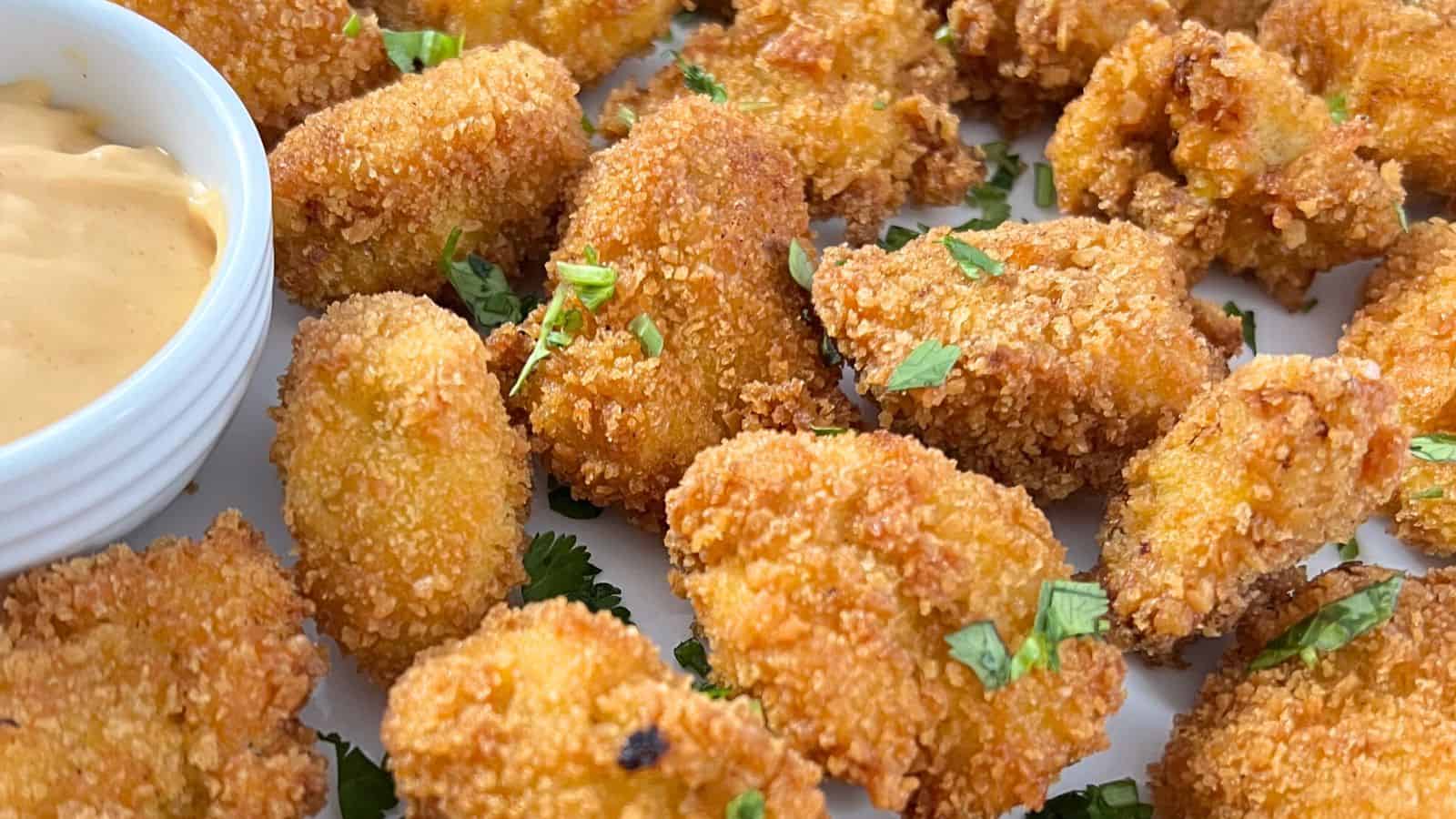Close-up of a plate of crispy, breaded fried chicken pieces garnished with chopped herbs, accompanied by a small bowl of dipping sauce on the side.