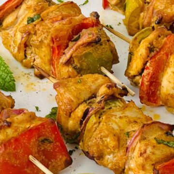 Close-up of grilled chicken kebabs on skewers with pieces of bell peppers, onions, and herbs, placed on a white plate garnished with mint leaves.