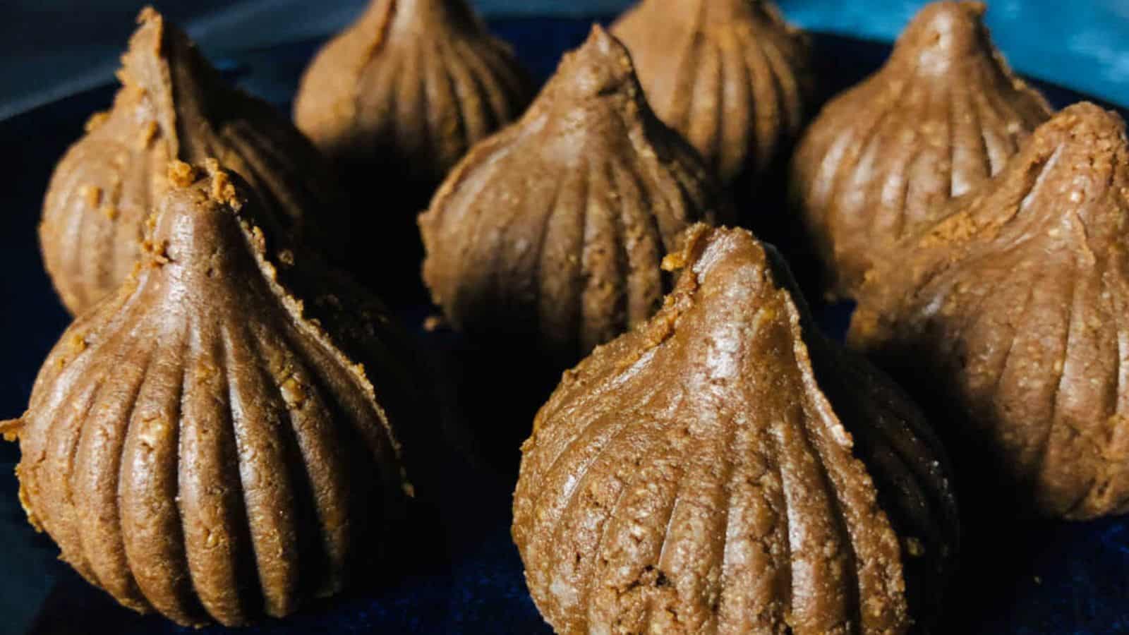 Close-up of eight modaks placed on a dark surface. These traditional Indian sweets have a ridged, conical shape and are made from brown dough.