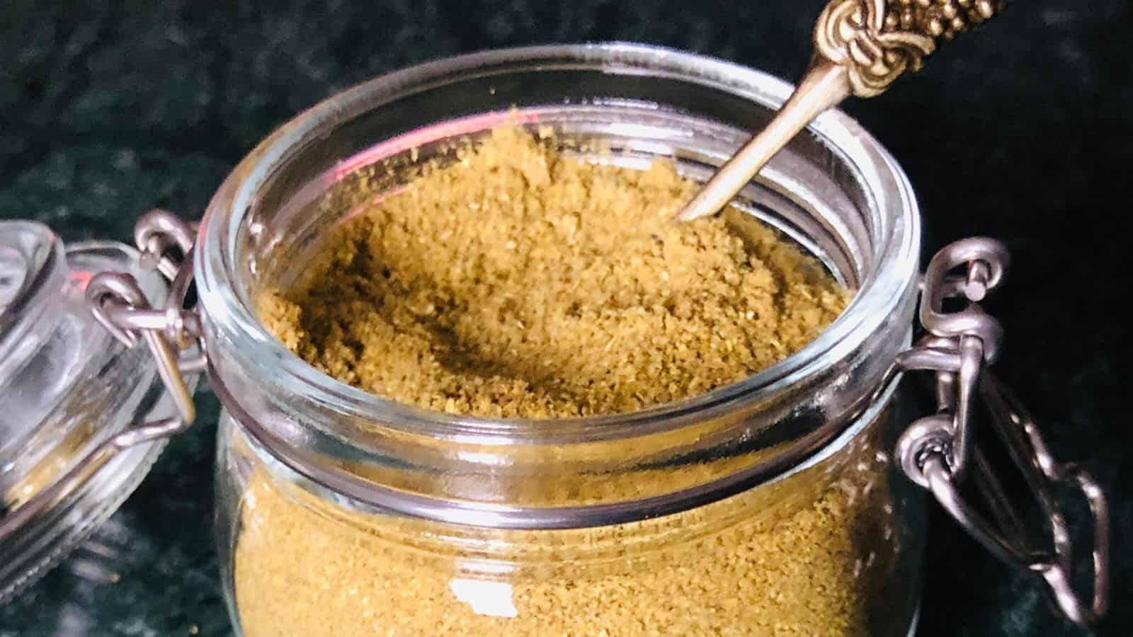 A clear glass jar with a hinged lid, filled with a yellowish-brown powder. A small golden spoon is partially submerged in the powder.