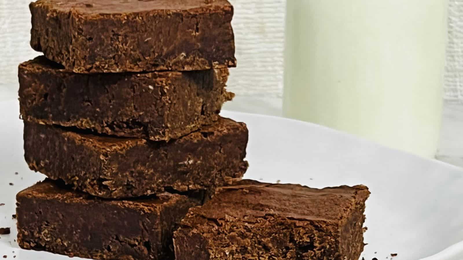 A stack of rich, fudgy brownies on a white plate, accompanied by a glass of milk in the background.