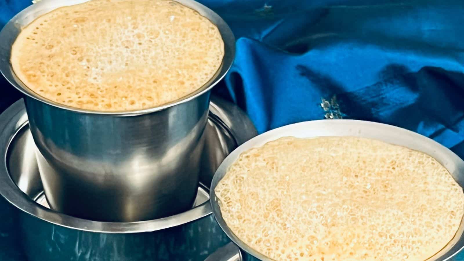Two containers of freshly made dosa batter on a blue cloth, one in a steel pot and another in a shallow dish overflowing with bubbles.