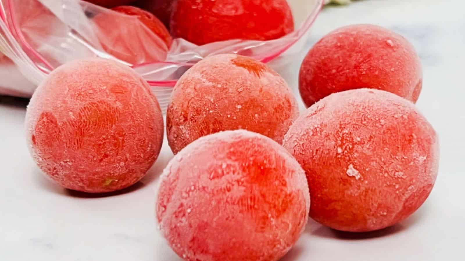 Five frozen tomatoes with a light frost coating, scattered on a surface, with some additional frozen tomatoes in a plastic bag in the background.