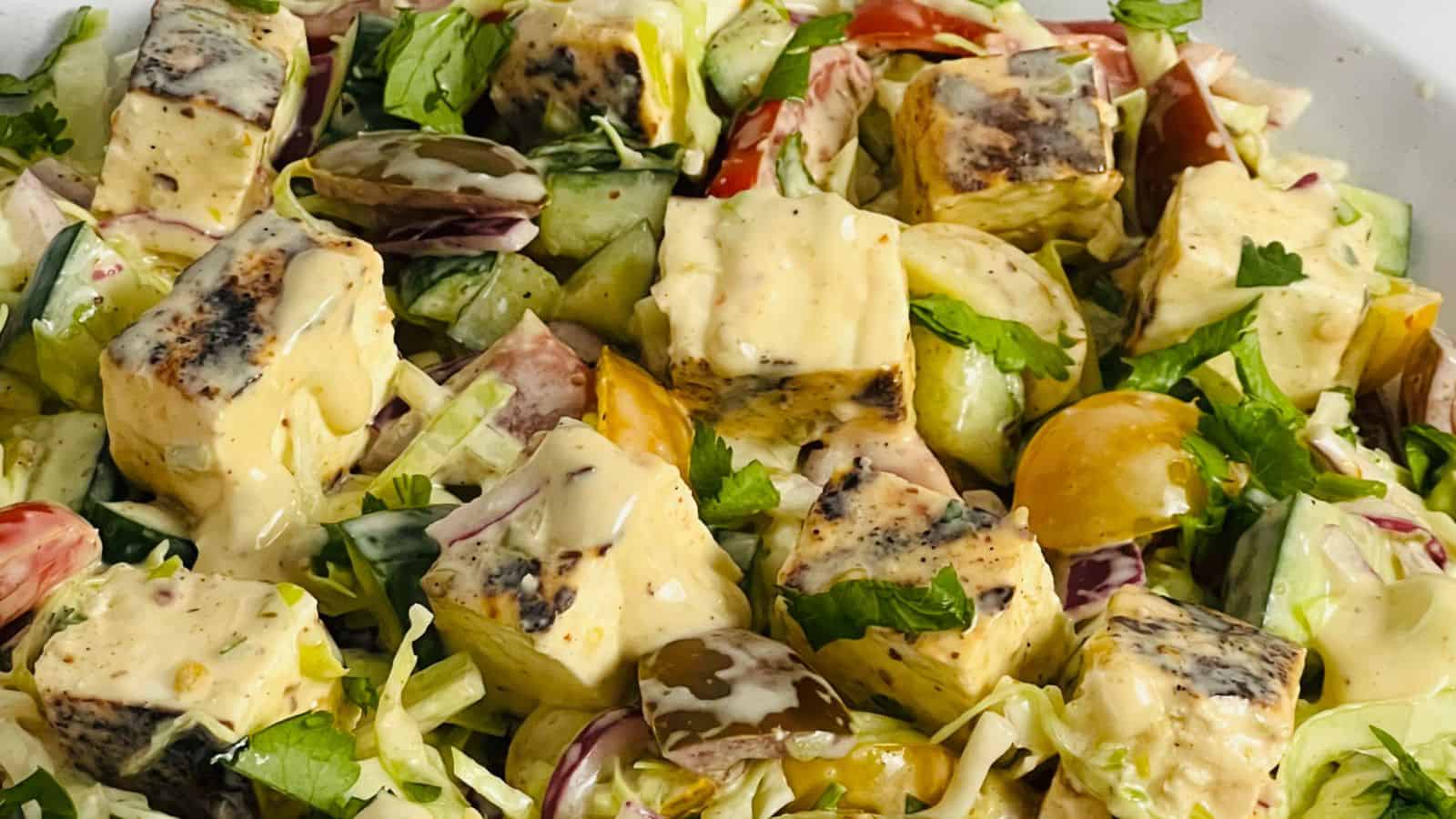 A close-up of a salad featuring grilled tofu cubes, cherry tomatoes, cucumber slices, and leafy greens, all drizzled with a creamy dressing.