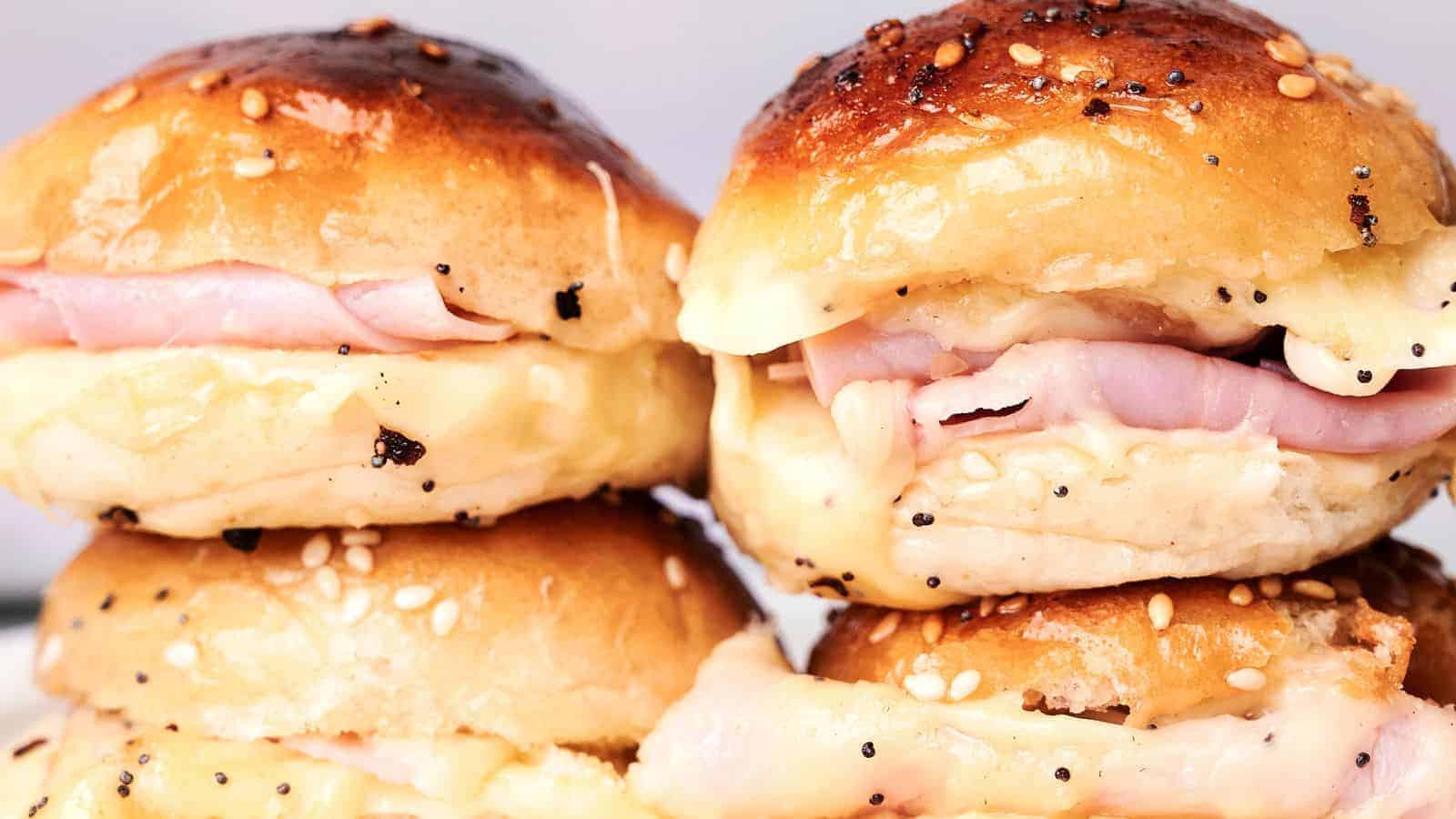 Close-up of four ham and cheese sliders on sesame seed buns, arranged in two stacks. The cheese is melting and oozing out of the sides, making these Ham and Cheese Sliders irresistible.