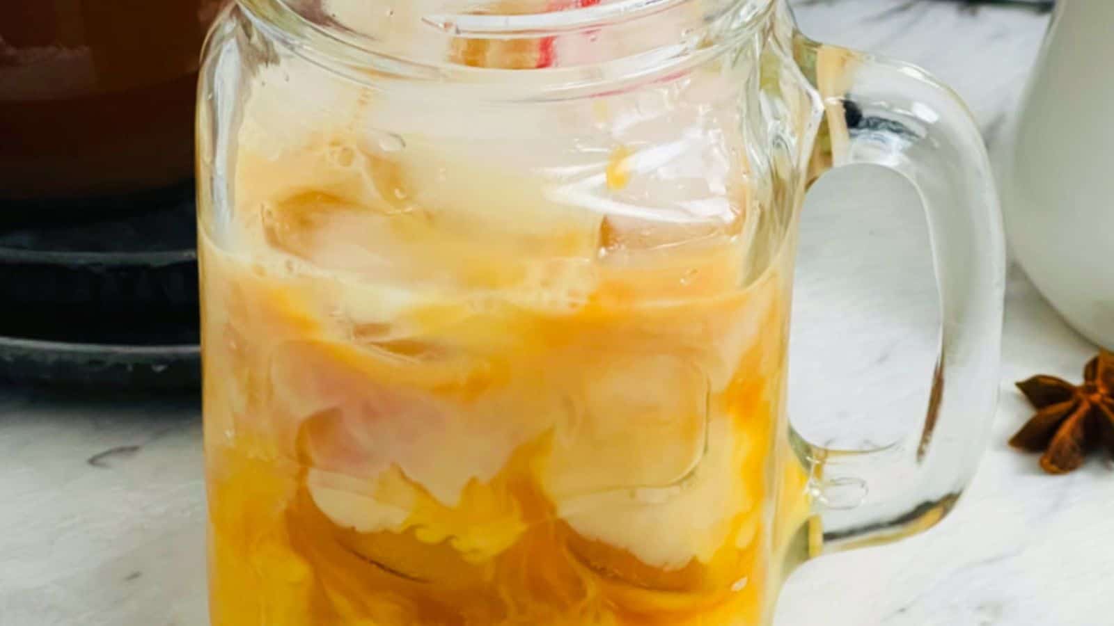 A glass mason jar filled with an iced beverage, likely an iced coffee or tea, with milk swirling into it.