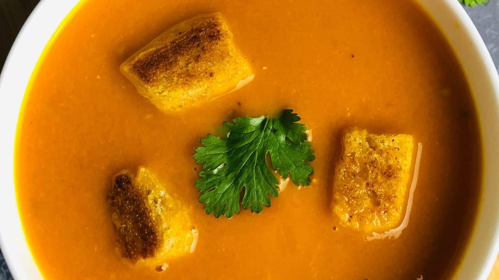 A bowl of orange soup with three golden-brown croutons and a sprig of parsley on top.