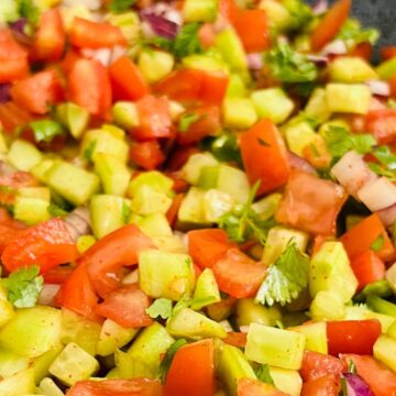 A close-up of a colorful diced vegetable mix, including tomatoes, cucumbers, red onions, and green herbs, in a gray dish.