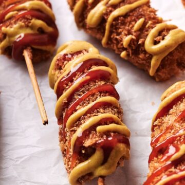 Several breaded and fried corn dogs on sticks, drizzled with mustard and ketchup, placed on white crumpled paper.