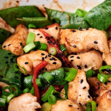 A close-up of a stir-fry dish featuring pieces of chicken, snap peas, red bell peppers, scallions, and black sesame seeds.
