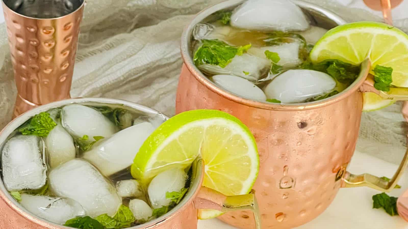 Two copper mugs are filled with iced drinks, garnished with lime slices and fresh mint leaves. A smaller copper cup is in the background.