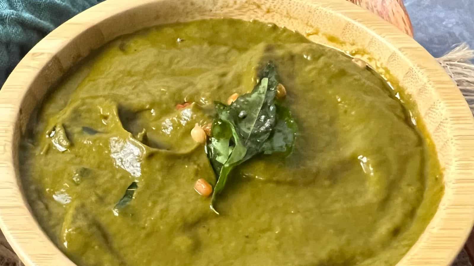 A bowl of green-colored curry garnished with coriander leaves and seeds. The curry is in a wooden bowl placed on a wooden surface.