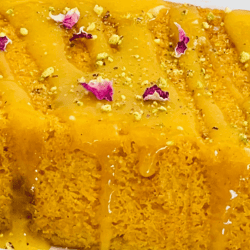Close-up of a vibrant orange dessert garnished with flower petals and nuts, drizzled with syrup on a white rectangular plate.